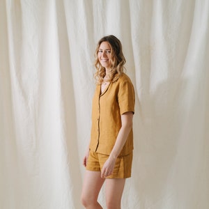 May include: A woman wearing a mustard yellow linen pajama set with a collared shirt and shorts. The set has a button-up front and short sleeves.