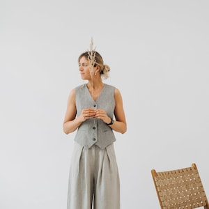 May include: A woman wearing a gray linen vest and matching wide-leg pants. She is holding a dried grass stalk in her hand and looking to the side. A wooden chair with a woven seat is in the background.