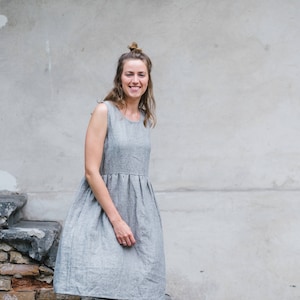 May include: A woman wearing a gray linen dress and brown sandals stands on a brick walkway in front of a gray wall.