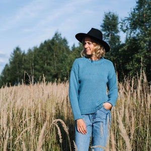 Pode incluir: Uma mulher vestindo um suéter azul e jeans está em pé em um campo de grama alta. Ela está usando um chapéu preto e olhando para o lado.