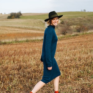 Op de afbeelding: Een vrouw in een tealblauwe trui-jurk en een zwarte fedora loopt door een veld met droog gras. Ze draagt ​​bruine laarzen en heeft een glimlach op haar gezicht.