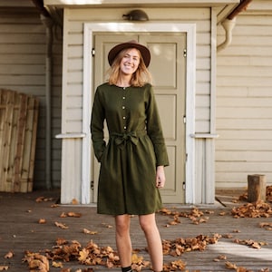 May include: A woman wearing a green linen dress with a brown hat and brown ankle boots stands in front of a wooden building with a red roof.  She is smiling and looking at the camera.  There are fallen leaves on the ground around her.