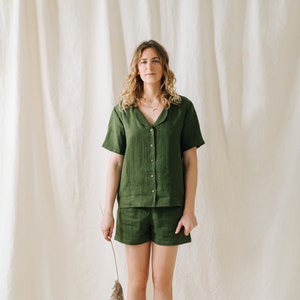 May include: A two-piece olive green linen pajama set. The set includes a short-sleeved button-down shirt and matching shorts. The model is holding a dried plant stem. The background is a neutral-colored fabric.