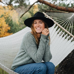 Puede incluir: Una mujer con un sombrero negro y un suéter verde se sienta en una hamaca blanca. Ella sonríe y mira a la cámara. La hamaca está colgada entre dos árboles.