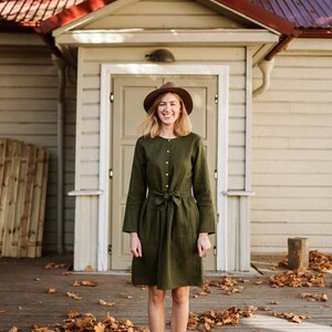May include: A woman wearing a green linen dress with a brown hat and brown boots stands in front of a house with a red roof. She is smiling and looking at the camera. There are fallen leaves on the ground around her.