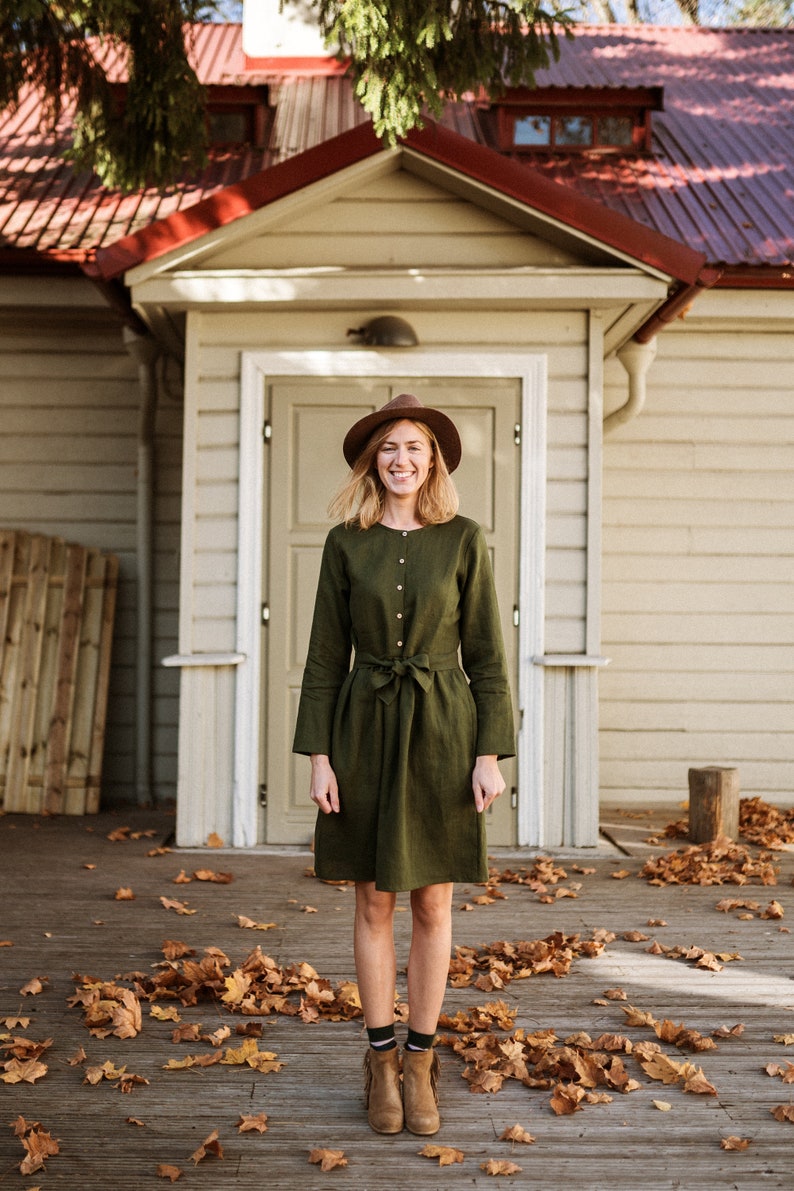 May include: A woman wearing a green linen dress with a brown hat and brown boots stands in front of a house with a red roof. She is smiling and looking at the camera. There are fallen leaves on the ground around her.