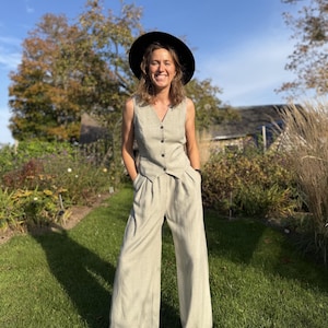 Op de afbeelding: Een lichtbeige mouwloos vest met een knoopsluiting aan de voorzijde en een bijpassende broek met hoge taille, wijde pijpen en zijzakken, gecombineerd met een zwarte hoed met brede rand en zwarte sandalen.