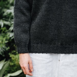 K&ouml;nnte beinhalten: Ein dunkelgrauer Strickpullover, der &uuml;ber einem wei&szlig;en Leinenhemd getragen wird. Der Pullover hat eine gerippte Struktur.