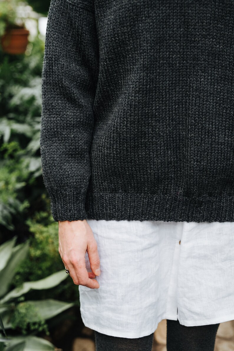 K&ouml;nnte beinhalten: Ein dunkelgrauer Strickpullover, der &uuml;ber einem wei&szlig;en Leinenhemd getragen wird. Der Pullover hat eine gerippte Struktur.