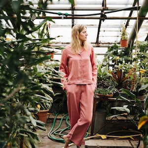 May include: A woman wearing a pink linen pajama set stands in a greenhouse. The set includes a long-sleeved shirt with a button-up front and matching pants.