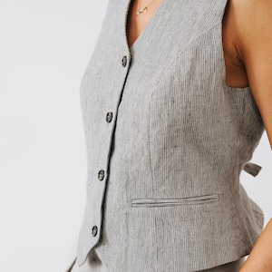 May include: A gray linen vest with a V-neckline and a single button closure. The vest has a pocket on the left side and a striped pattern.