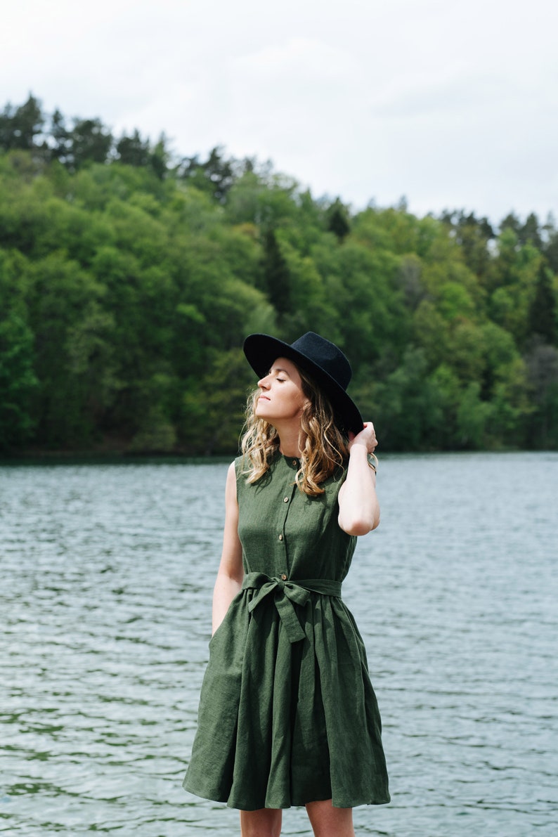 May include: A woman wearing a green linen dress with a black hat stands by a lake. The dress has a belt and pockets. The lake is surrounded by green trees.