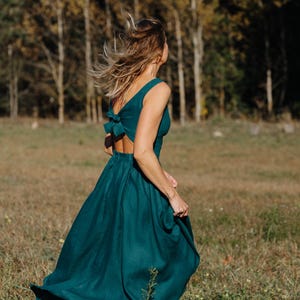 Peut inclure: Une robe fluide de couleur sarcelle avec un d&eacute;tail de n&oelig;ud dans le dos. La robe sans manches a une jupe ample et est port&eacute;e &agrave; l'ext&eacute;rieur dans un champ avec des arbres en arri&egrave;re-plan. La robe semble &ecirc;tre en lin ou dans un tissu similaire.