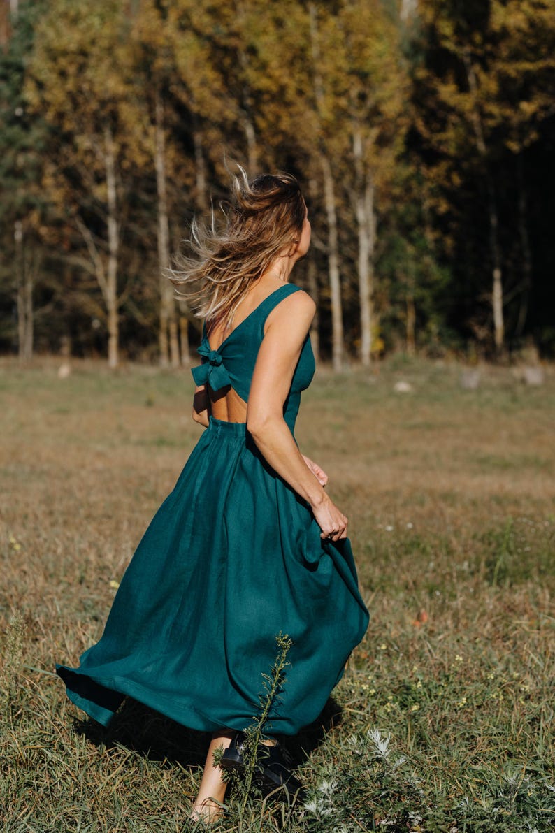 Peut inclure: Une robe fluide de couleur sarcelle avec un d&eacute;tail de n&oelig;ud dans le dos. La robe sans manches a une jupe ample et est port&eacute;e &agrave; l'ext&eacute;rieur dans un champ avec des arbres en arri&egrave;re-plan. La robe semble &ecirc;tre en lin ou dans un tissu similaire.