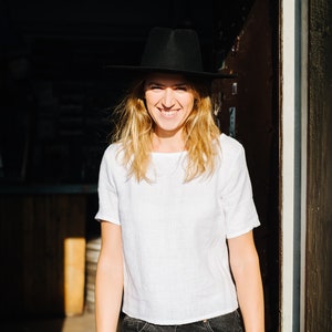 Op de afbeelding: Een vrouw draagt een wit linnen shirt, een zwarte jeans en een zwarte fedora-hoed en glimlacht naar de camera. Ze staat in een deuropening met zonlicht dat op haar schijnt.