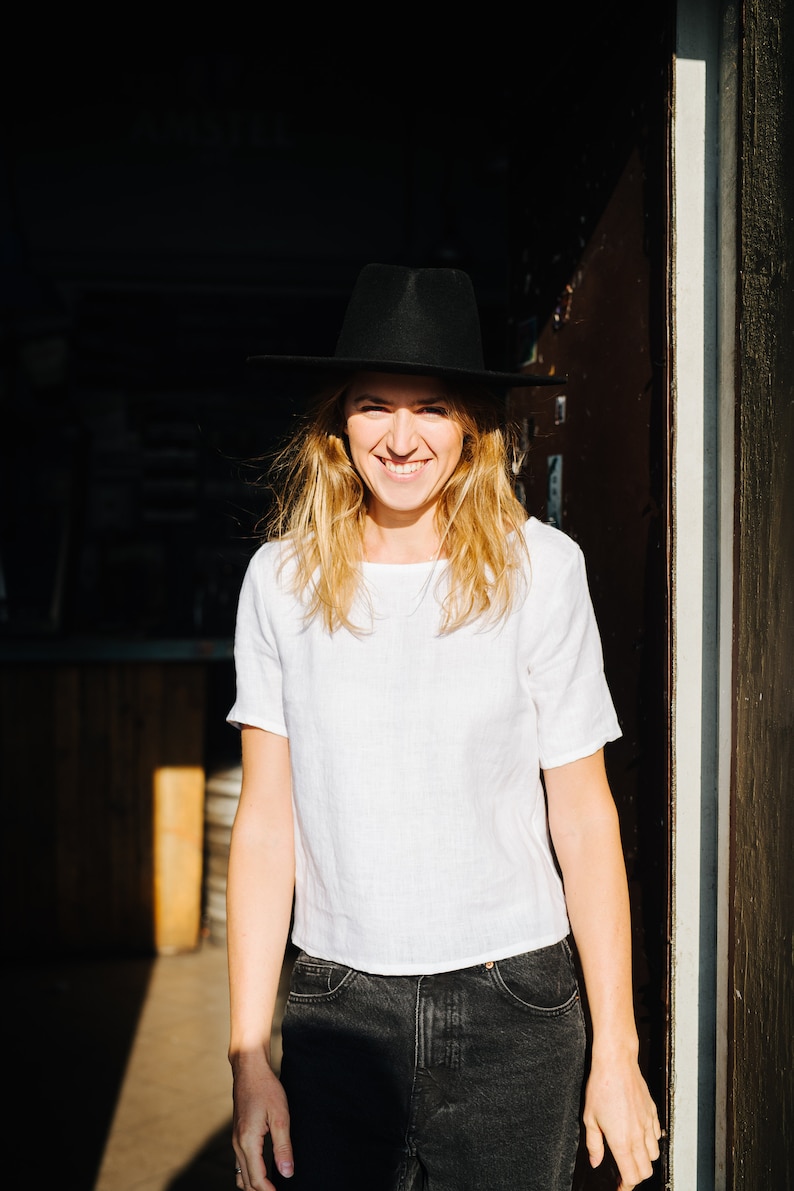 Op de afbeelding: Een vrouw draagt een wit linnen shirt, een zwarte jeans en een zwarte fedora-hoed en glimlacht naar de camera. Ze staat in een deuropening met zonlicht dat op haar schijnt.