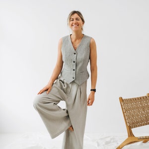 May include: A woman wearing a gray vest and wide-legged pants, standing on one leg with a relaxed smile.