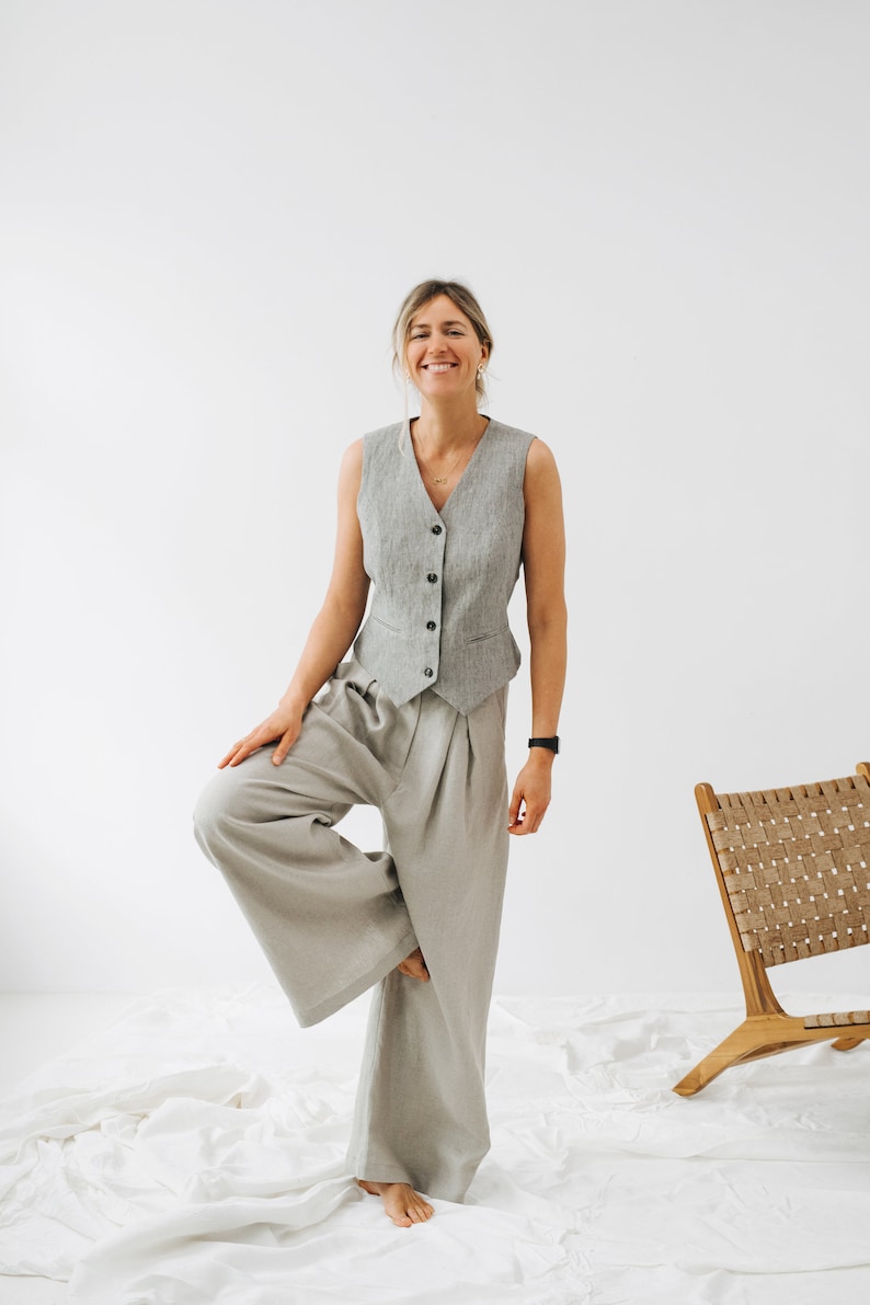May include: A woman wearing a gray vest and wide-legged pants, standing on one leg with a relaxed smile.