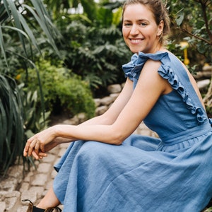 Puede incluir: Una mujer con un vestido de lino azul claro con volantes est&aacute; sentada en un camino de piedra en un jard&iacute;n. Ella sonr&iacute;e y mira a la c&aacute;mara.