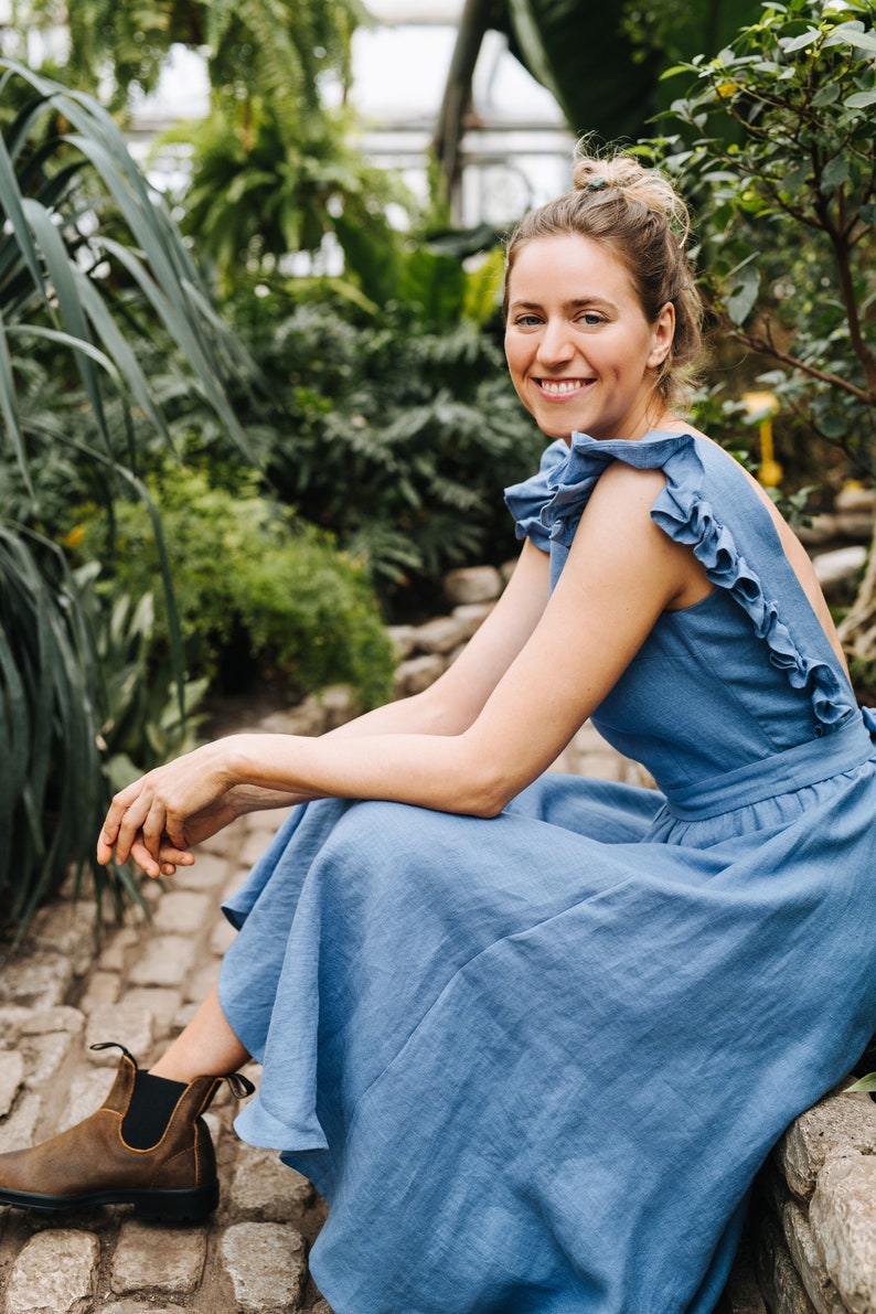 Puede incluir: Una mujer con un vestido de lino azul claro con volantes est&aacute; sentada en un camino de piedra en un jard&iacute;n. Ella sonr&iacute;e y mira a la c&aacute;mara.