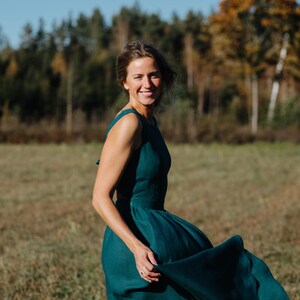 Peut inclure: Une robe sans manches de couleur teal avec une jupe fluide. La robe est faite d'un tissu doux et a un corsage ajust&eacute;. La personne portant la robe sourit dans un champ avec des arbres en arri&egrave;re-plan.