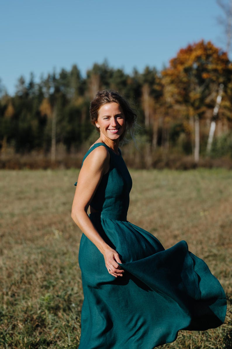 Peut inclure: Une robe sans manches de couleur teal avec une jupe fluide. La robe est faite d'un tissu doux et a un corsage ajust&eacute;. La personne portant la robe sourit dans un champ avec des arbres en arri&egrave;re-plan.