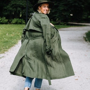 May include: A woman wearing a green linen trench coat walks down a path in a park. She is wearing a hat and jeans.