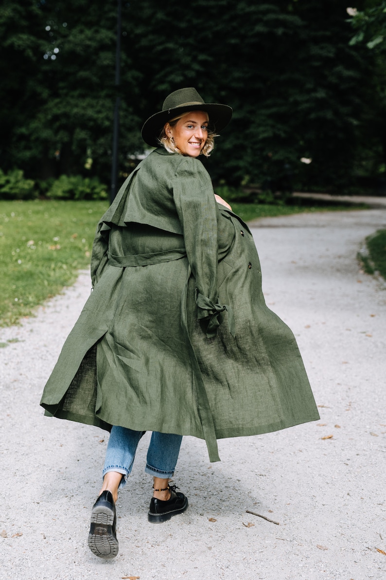 May include: A woman wearing a green linen trench coat walks down a path in a park. She is wearing a hat and jeans.
