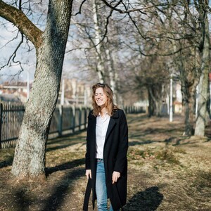 May include: A woman in a black coat and blue jeans walks through a park with bare trees. She is smiling and looking to the side.
