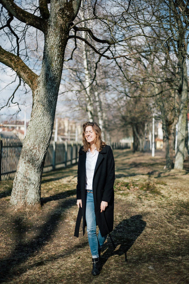 May include: A woman in a black coat and blue jeans walks through a park with bare trees. She is smiling and looking to the side.