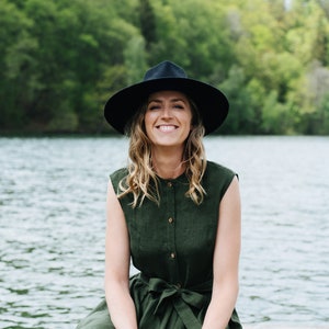 May include: A woman wearing a green linen dress with a black hat sits on a wooden dock by a lake. She is smiling and looking at the camera.