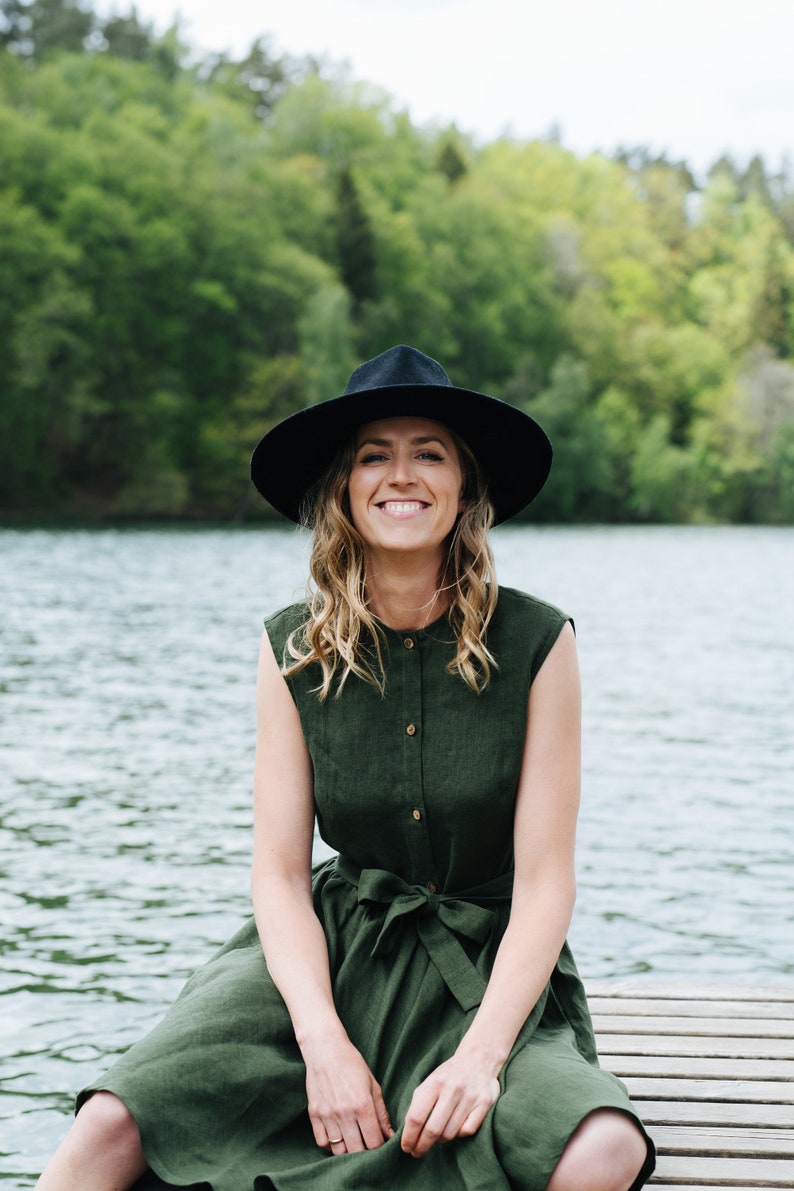 May include: A woman wearing a green linen dress with a black hat sits on a wooden dock by a lake. She is smiling and looking at the camera.