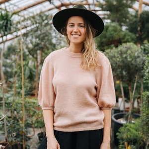 May include: A woman wearing a pink sweater and a black skirt stands in a greenhouse. She is wearing a black hat and smiling at the camera.