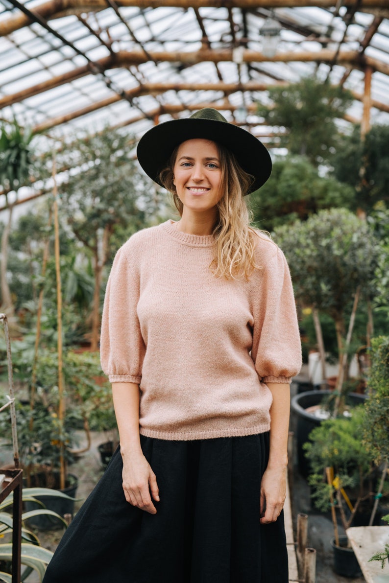 May include: A woman wearing a pink sweater and a black skirt stands in a greenhouse. She is wearing a black hat and smiling at the camera.