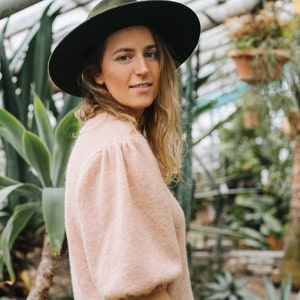 May include: A woman wearing a light pink sweater with puffed sleeves and a dark green fedora hat. She is standing in a greenhouse with lush green plants in the background.