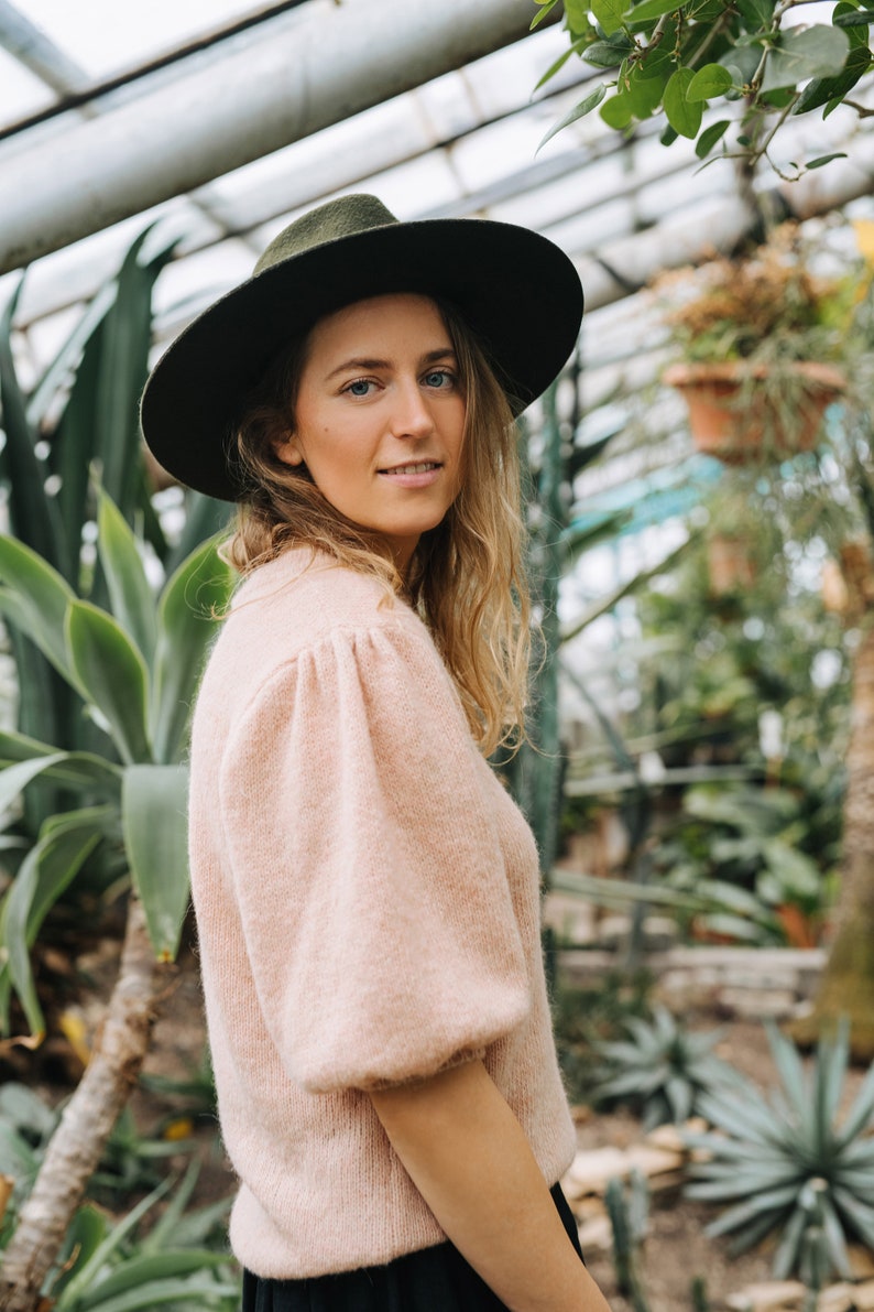 May include: A woman wearing a light pink sweater with puffed sleeves and a dark green fedora hat. She is standing in a greenhouse with lush green plants in the background.