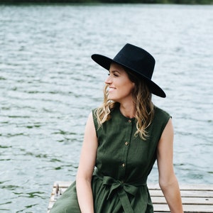 May include: A woman wearing a green dress and a black hat sits on a wooden dock overlooking a lake.