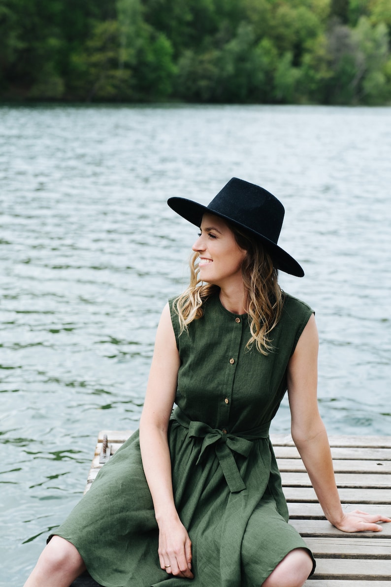 May include: A woman wearing a green dress and a black hat sits on a wooden dock overlooking a lake.