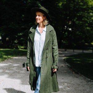 May include: A woman wearing a green linen trench coat, a wide-brimmed hat, and blue jeans walks on a gravel path.