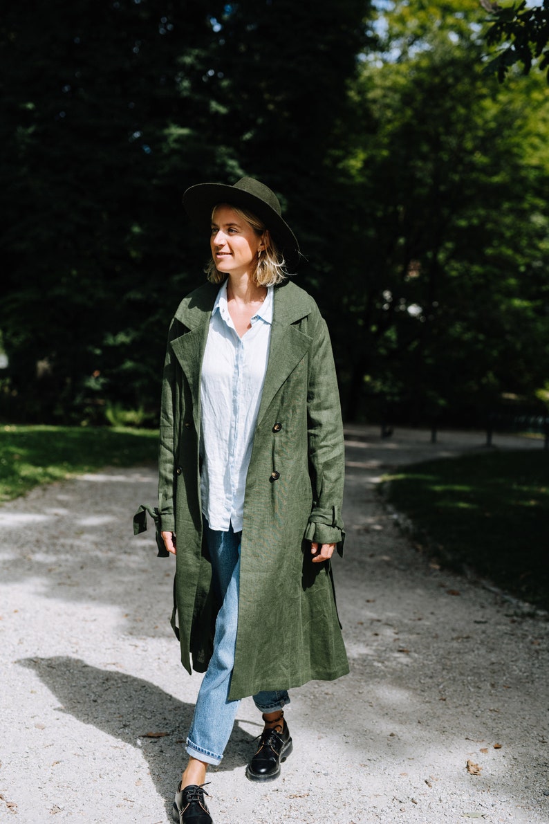 May include: A woman wearing a green linen trench coat, a wide-brimmed hat, and blue jeans walks on a gravel path.
