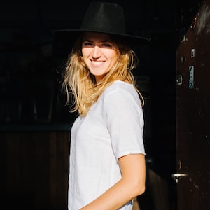 Op de afbeelding: Een vrouw draagt een zwarte hoed, een wit T-shirt en een donkere spijkerbroek. Ze glimlacht en kijkt naar de zijkant.