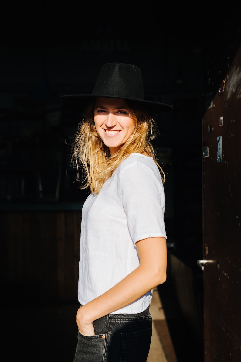 Op de afbeelding: Een vrouw draagt een zwarte hoed, een wit T-shirt en een donkere spijkerbroek. Ze glimlacht en kijkt naar de zijkant.