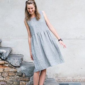 May include: A woman wearing a gray linen dress and brown sandals stands in front of a stone wall. She is smiling and looking down.