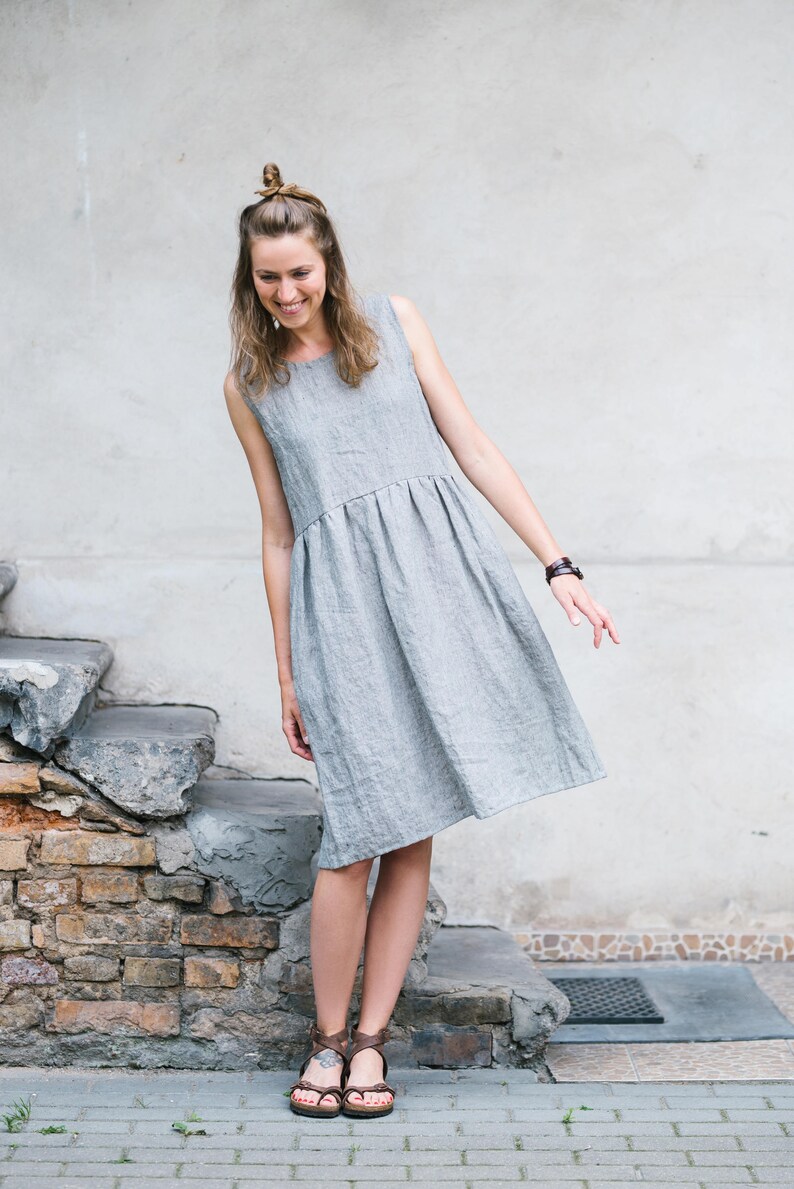 May include: A woman wearing a gray linen dress and brown sandals stands in front of a stone wall. She is smiling and looking down.