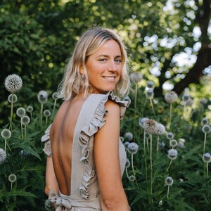 May include: A woman with blonde hair is wearing a light brown linen dress with a low back and ruffles on the shoulders. She is standing in a garden with green plants and blue flowers.