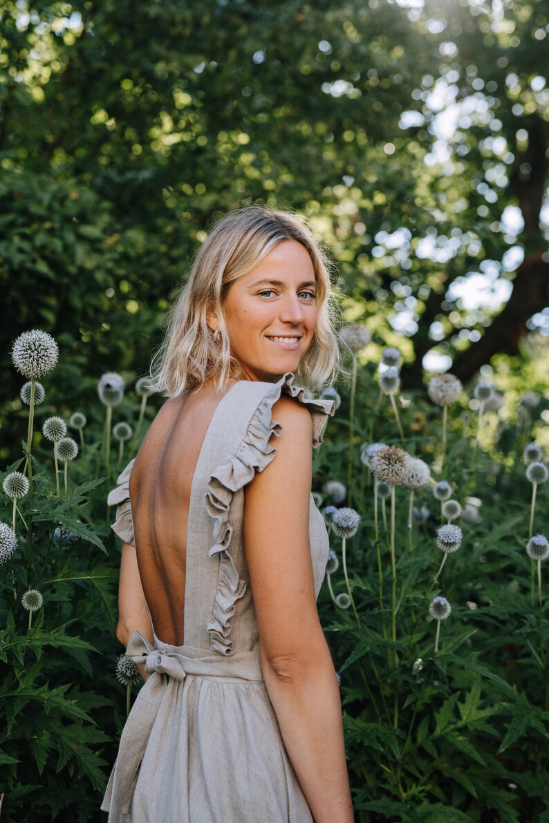 May include: A woman with blonde hair is wearing a light brown linen dress with a low back and ruffles on the shoulders. She is standing in a garden with green plants and blue flowers.