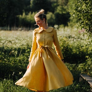 Pode incluir: Uma mulher com um vestido de linho amarelo com colarinho de babados e cintura amarrada est&aacute; em um campo de flores silvestres.