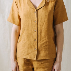 May include: Mustard-colored linen pajama set. The short-sleeved button-down shirt has a notched collar and wooden buttons. The matching shorts complete the set. The fabric has a textured appearance.