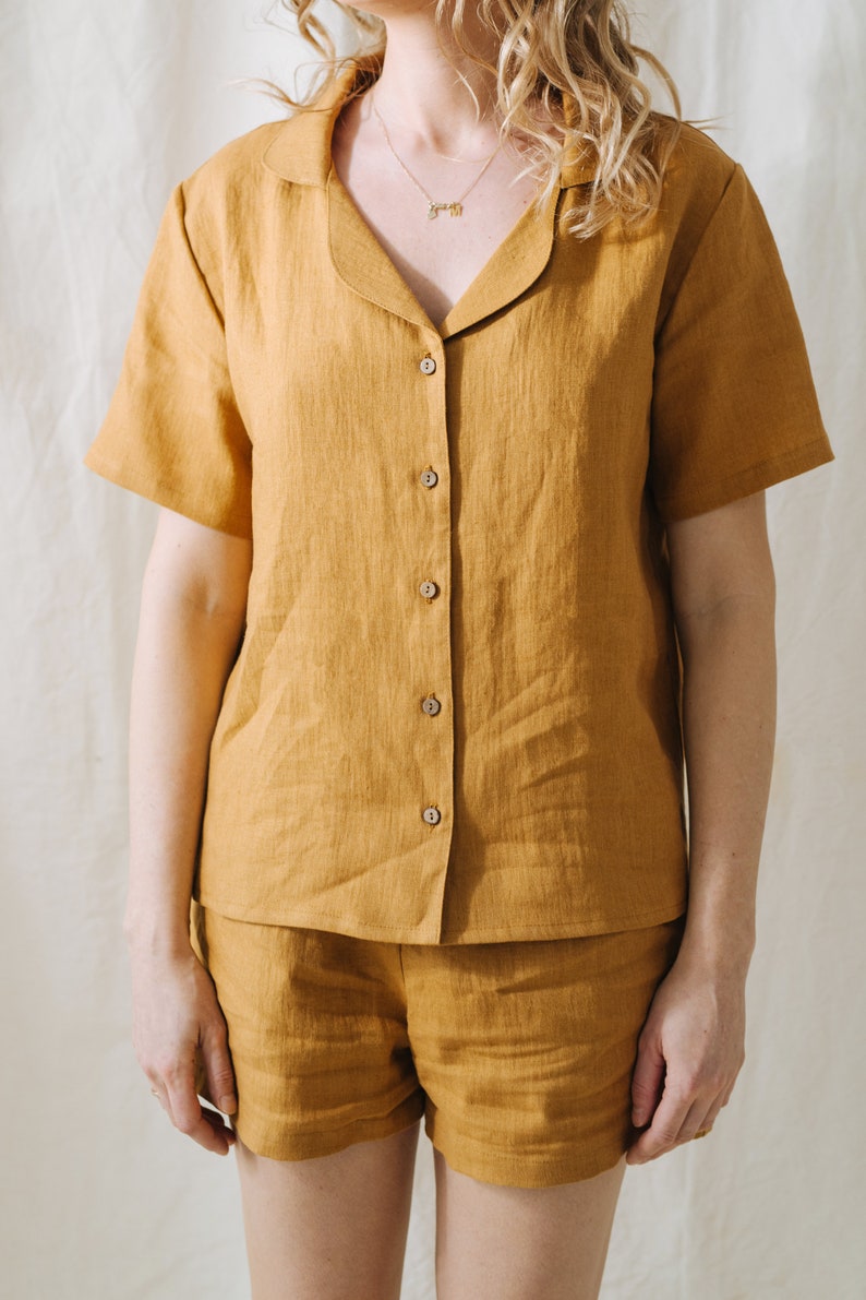May include: Mustard-colored linen pajama set. The short-sleeved button-down shirt has a notched collar and wooden buttons. The matching shorts complete the set. The fabric has a textured appearance.