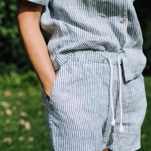 May include: A two-piece linen outfit in a vertical striped pattern of white and green. The outfit includes a short-sleeved button-down shirt and drawstring shorts. The shorts have pockets and a white drawstring. The outfit is made of a lightweight fabric.